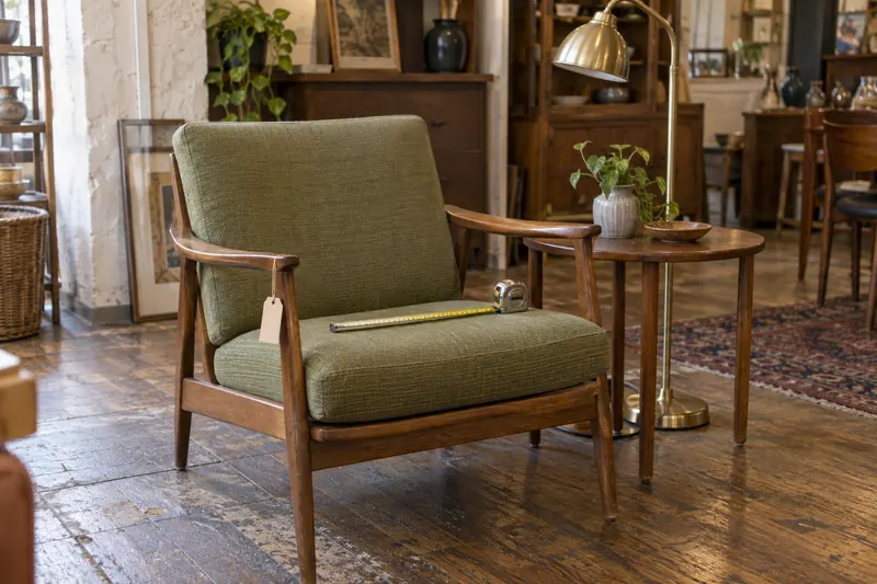 A mid-century armchair with green cushions and a tape measure on the seat, next to a round side table and a brass floor lamp